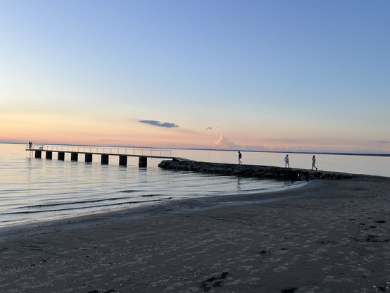 Bathing pier leading into the calm water of the Baltic Sea. The sky is tinged yellow and orange near the horizon and a few clouds are visible in the distance. Three kids are slowly walking onto the pier.