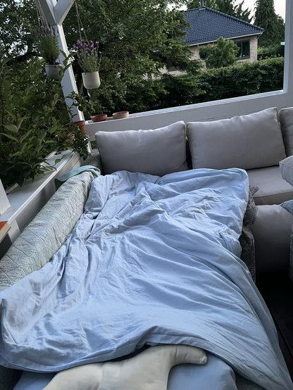 Terrassesofa dækket med vattæppe, dyne og puder. I baggrunden kig ud til grønne planter