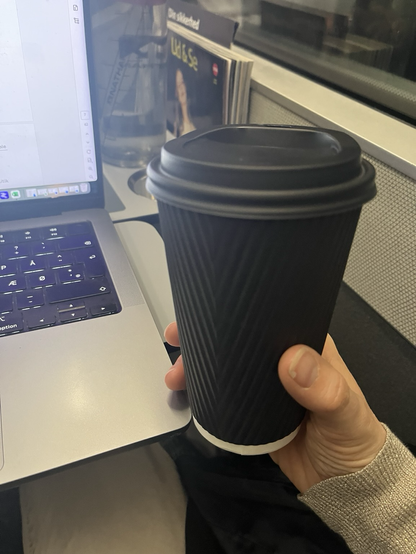 Sort engangskrus med kaffe holdt af en højrehånd. Til venstre i billedet lidt af en opslået bærbar computer på et DSB-togbord