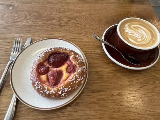 Cafébord med tallerken med en slags briochebolle med ferskencreme og udskårne jordbær. Til højre en kop cappuccino med fint mælkemønster