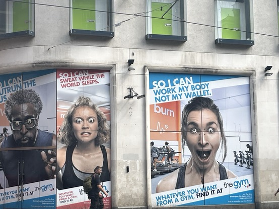 Store butiksvinduer der er dækket til med plakater for et eller andet fitness-studie (?). Der er tre personer i halvnær beskæring hvilket gør deres hoveder mindst et par meter høje. På alle tres øjne er sat googly eyes i den rigtige (store!) størrelse