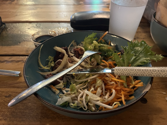Træbord med en skål blandet vietnamesisk med kniv og gaffel liggende på tværs. Bagved et glas lemonade og et brilleetui