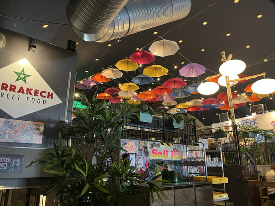 Til venstre et skilt med ordlyden Marrakech Street Food. Midtfor og hen til højre en masse kulørte paraplyer ophængt under det sorte loft i en stor sal. Derudover grønne planter og overflader med graffiti
