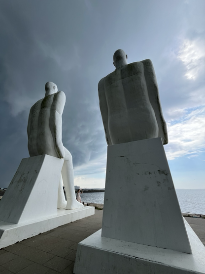 Nærbillede af to af skulpturerne bagfra med kig ud til lyseblå himmel ud over havet og truende mørke skyer over deres hoveder