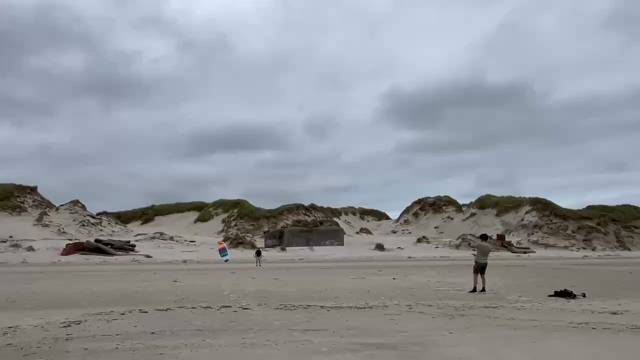 Med ryggen til Vesterhavet ses ÆldsteSøn styre en regnbuefarvet drage i buer og cirkler op og ned. I baggrunden bunkere og klitter og en noget grå himmel