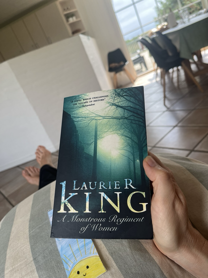 Laurie R. Kings roman A Monstrous Regiment of Women, forsiden holdt i toner af grøn og sort, holdt op over en stribet sofapude. På puden ligger også et hjemmelavet bogmærke med en gul, håndtegnet sol på en lyseblå himmel med hvide skyer 