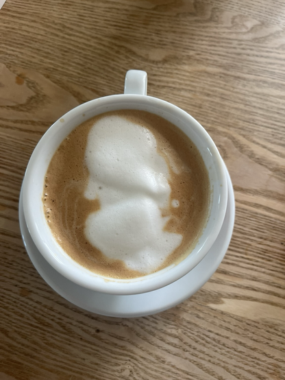 Hvid kaffekop på træbordplade. Billedet er taget direkte ovenfra så man kan se den kanelbrune kaffe i midten af hvilken ligger mælkeskum der omtrent ligner en russer fra tsar-tiden