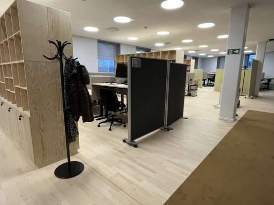 Storrumskontor opdelt af høje birketræsreoler og mørkegrå, flytbare skærme. Man kan se en brunlig løber (tæppe, altså) på gulvet, et par hvide søjler der holder loftet og en masse alt for skarpt lys. Inde bag den ene grå skærm en række skriveborde hvoraf det første er kørt op i stå-position. Foran den nærmeste reol står en stumtjener med en jakke og et tørklæde på