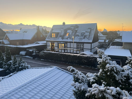 Udsigt fra vindue på første sal over en villavej med huse og træer dækket af et let lag sne. Solen står op i baggrunden og gør himlen gul og orange
