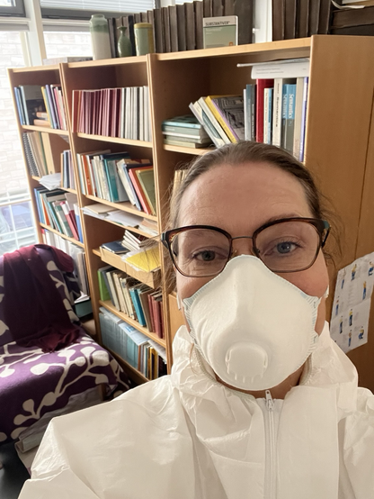 Overtegnede iført åndedrætsværn og sikkerhedsdragt foran en reol fyldt med bøger og papirer