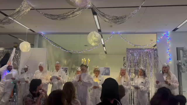 Video der viser otte personer iklædt hvide beskyttelsesdragter synger julen ind. I midten en kvinde med lyskrans om hovedet og hænderne i bedegestus foran brystet. I loftet både sølvglitter og kaffefilterlamper. Foran kolleger der kigger med