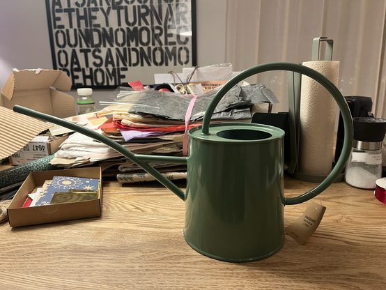 Vandkande i grøn metal med stor buet håndtag og en lang, tynd tud. Placeret på et træbord foran en masse blandet gavepapir og kort. Man aner noget bølgepap til den ene side (klogt!)