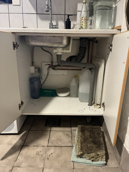 Bryggersvask med åbne skabslåger ind til rør og rod. På gulvet foran et sammenfoldet håndklæde med en halv fortovsflise på eller noget??)