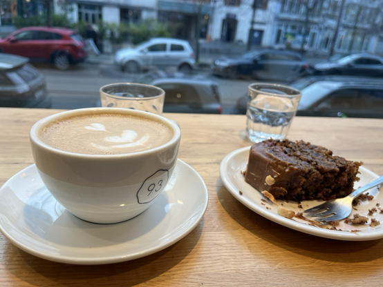 En stor kop cappuccino på et træbord med udsigt til gaden. Ved siden af en halvt spist kage med chokolade, kirsebær og karamel - mums!)