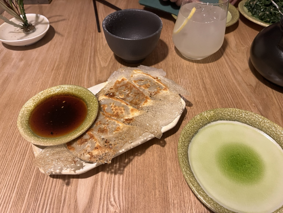 Udsnit af servering med forretter og drikkevarer. Til venstre gyoza stegt så de hænger sammen, ved siden af en lav, bred skål soya. Til højre en mindre, tom tallerken i grønne nuancer. I baggrunden en skål til tesien, et glas yuzu limonade og en kande sencha-te