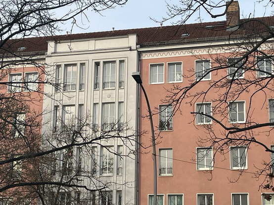 Husfacade i støvet, lys rød/mørk laksefarvet med hvide rammer om vinduerne og er bånd under taget med en rød, bugtet linje. Midt på facaden en sektion i lys grå/hvid som er fremhævet fra resten og har høje, smalle vinduer