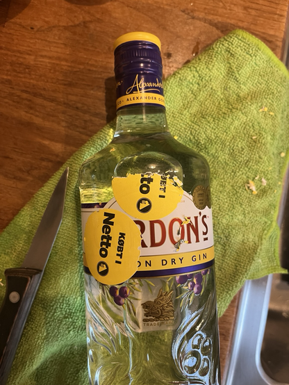 En flaske Gordon’s gin liggende på en grøn karklud med en kniv ved siden af. Det ene af to reklamegule klistermærker med teksten “Købt i Netto” er delvist skrabet af, og der ses papirfnuller rundt omkring som vidne om besværligheden