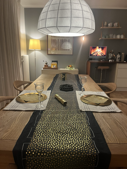 Nytårsbord dækket til to med gyldne undertallerkener, en sort løber med en ekstra løber ovenpå med guldpletter. I midten én gylden knallert og i baggrunden tre små bordbomber