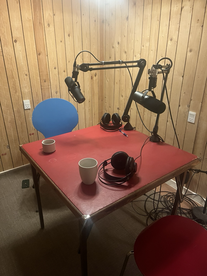 Lille rødt bord i et rum med trævægge. Der er hørebøffer og mikrofoner og kaffekopper klar plus en fin blå stol