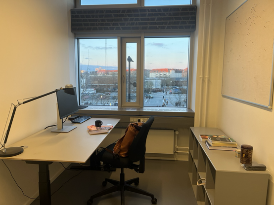 Kontor med skrivebord til den ene side, vinduesparti for enden og to lave reoler og en opslagstavle til den anden side. Uden for vinduet udsigt over Islands Brygge. På toppen af en lygtepæl sidder en krage og kigger ud på verden