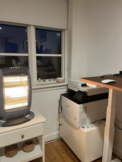 Hjemmekontor hvor man kan se lidt af et hævesænkebord og en printer til højre, et vinduesparti midt for, og en tændt varmeovn til venstre. Den udsender lys i bræmmer. Himlen udenfor er stadig ret mørk