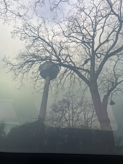Billede taget gennem taget på en bil. Det hele fremstår tåget. Motivet er et Fernsehturm (?) i Köln. I forgrunden et træ og en gadelygte. Genskin inde fra bilen af vinduerne på bag- og forsæde