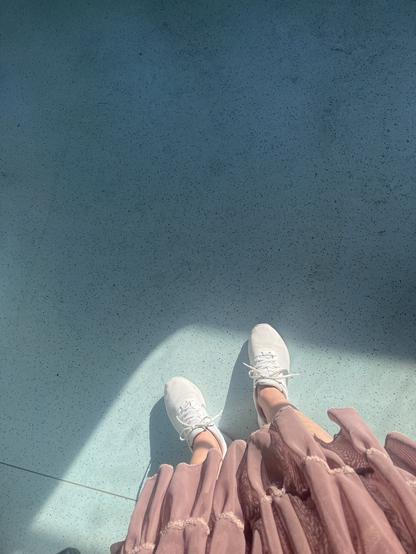 Billede taget oppefra af gammelrosa tylskørt med et par blege sommerben stikkende ud, afsluttet af hvide sneakers. Fødder og resten er placeret i en metro hvor solen skinner ind og indrammer et felt omkring skoene