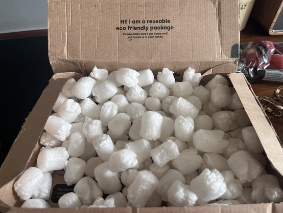 Åben papkasse til varmelevering, fyldt med hvide, skumagtige, korte “pølser”. I låget på pakken står der: Hi! I am a reusable eco friendly package