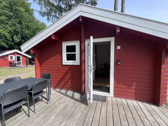 Rødmalet træhytte med lille terrasse og udendørs møbler. Døren står på vid gab og solen skinner 
