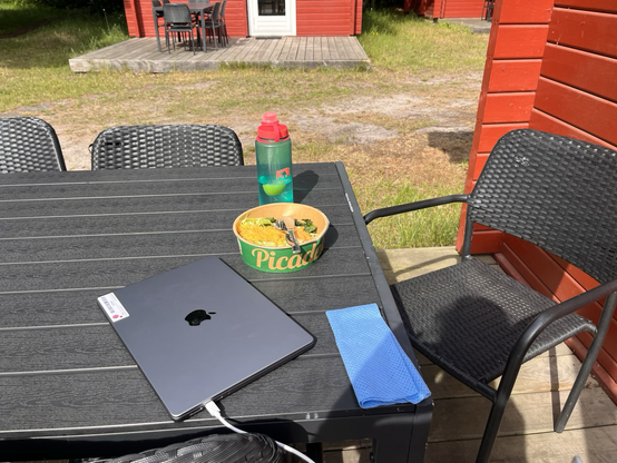 Udendørs bord og stole på terrassen foran hytten. På bordet står en drikkedunk, en bøtte med blandet salat, en computer og en lille, blå klud (her er ingen køkkenrulle)