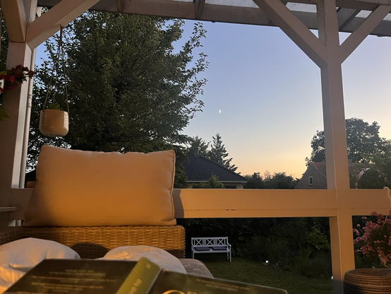 Aftenudsigt fra sofaen på terrassen. Himlen changerer mellem lys blålilla og pastelgul. Månen hænger med et fint sejl ved siden af det store grantræ. Det hele er indrammet af terrassens hvidmalede bjælker