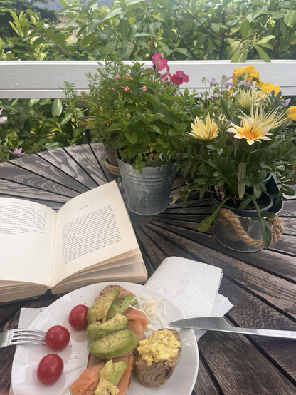 Rundt havebord med krukker med blomster og krydderurter. På bordet ligger en opslået bog bag en tallerken med en laksemad med avocado og en lille bid brød med æggesalat. Dertil tre tomater og lidt peberrod