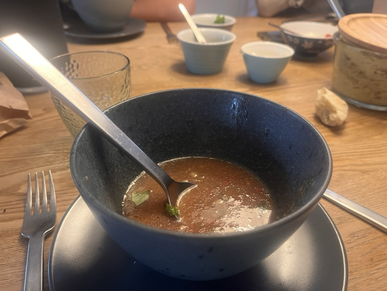 Grå keramikskål med gazpacho og en ske. Man kan se skåle i baggrunden til de finthakkede grøntsager der skal på toppen