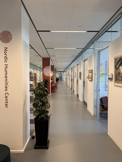 Lang kontorgang med bredt midterparti inddelt af søjler og planter. På den nærmeste søjle står der Nordic Humanities Center nedad. Man kan se én åben dør ind til et kontor hvor man kan skimte en lænestol og nogle post-its på væggen. Der er kunst på væggene mellem flere af kontorerne (min fars, sagde hunden!)