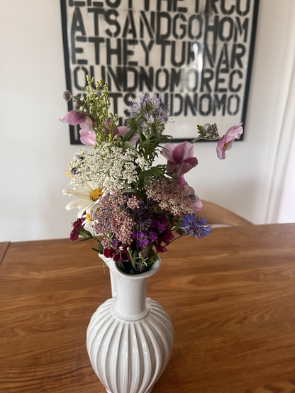 I en hvid keramikkande er sat en lille buket blandede blomster, blandt andet vild gulerod, msrguerit, høstanemone, kornblomster, en snerle, et par nelliker og en honningurt. Farverne er hvid, blå, lyserød, lilla og gul