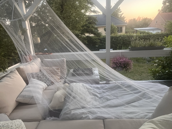 En oppustelig madras med dyne og puder placeret på en terrasse foran en hjørnesofa og med et myggenet bredt ud over hele molevitten. I baggrunden udsigt til grøn have og lidt lyserøde blomster