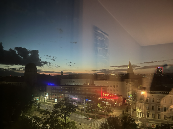 Udsigt fra hotelværelse på 7. etage ud over Szczecin, Polen. Aftenhimlen er mørk øverst men changerer i lysere blå, gul og orange ned mod horisonten. Kirketårne stikker op til højre og venstre, det ene foran en klump mørke skyer. Nede foran diverse etagebyggerier er en åben plads og en bred allé. Der er enkelte, store træer også. Venstre del af billedet er sløret af et gulligt lys fra værelset og man aner et kunstværk på væggen og linjen mellem væg og loft. Helt til højre ses skyggen af den hån…
