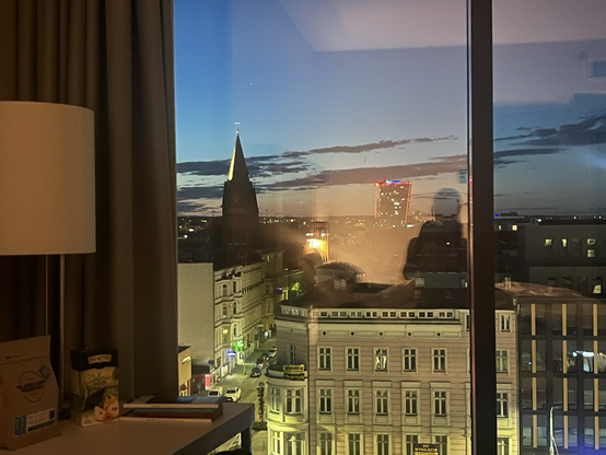 Lignende motiv som før, men her ser man mere af hotelværelset, nemlig en lampe og et gardin til højre og en ramme midt i det store gulv-til-loft-vindue. Udsigten viser et kirketårn til venstre og en smuk, ældre etageejendom midtfor. Andre, mere moderne, byggerier til højre Aftenhimlen fremstår lysere med mørke skyer i en længere stribe fra venstre til højre. Solnedgangen smelter sammen med det gule lys inde fra værelset. Skyggen af en kvinde der holder et kamera op foran sig kan ses i det gule …