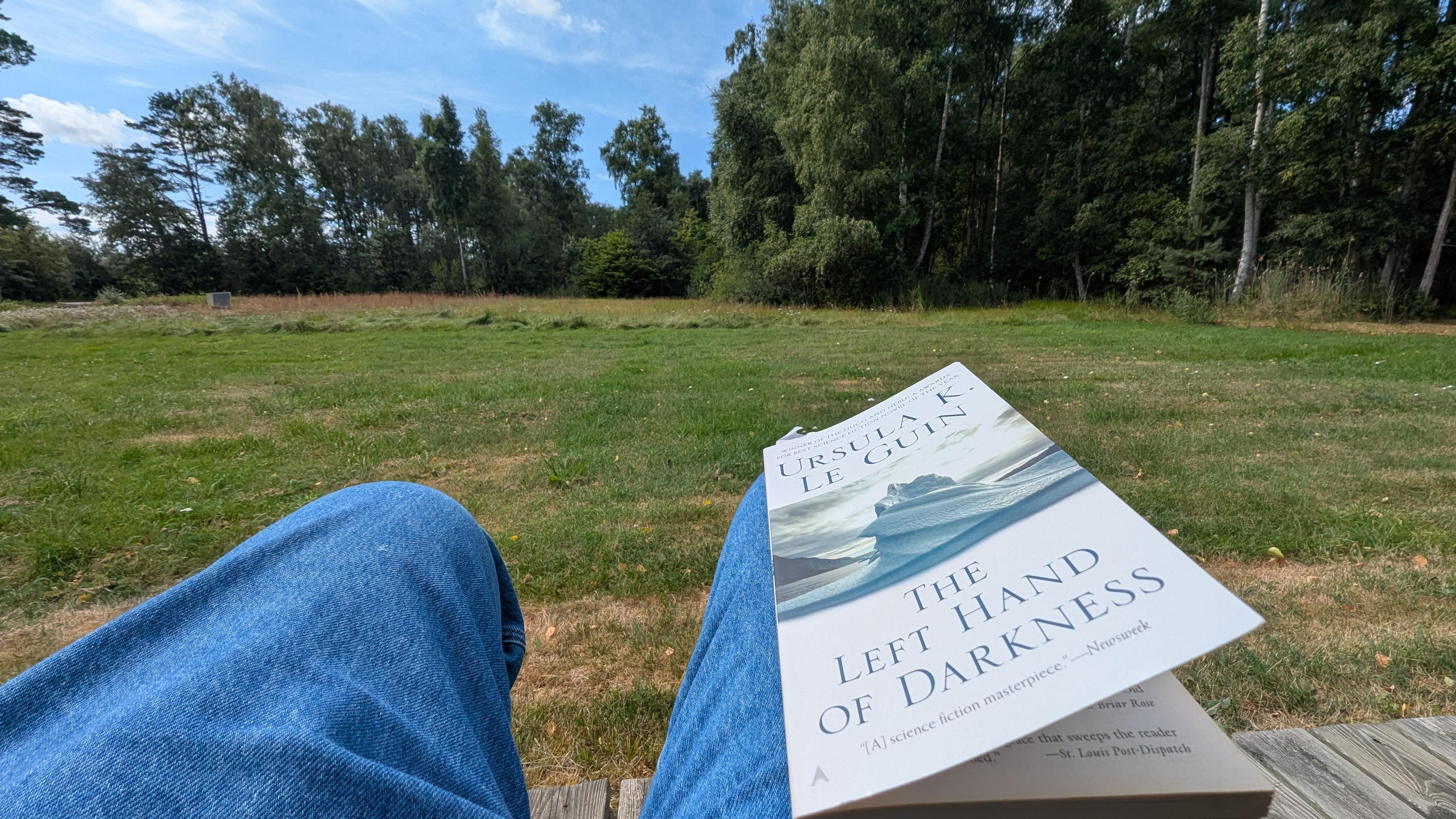 Udsigt mod stor have med høje træer i baggrunden. I forgrunden 2 ben. På det ene ligger min bog: Ursula K. Le Guin: the left hand of Darkness