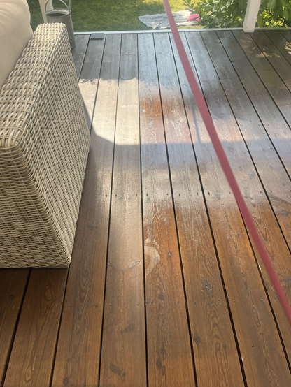 Træterrasse hvor en sektion til højre tydeligt er blevet olieret og resten ikke er. Det olierede er sådan nogenlunde afmærket med en snor (bordeaux gavebånd). Et fodaftryk ses i det blanke olierede. Til venstre lidt af en havesofa