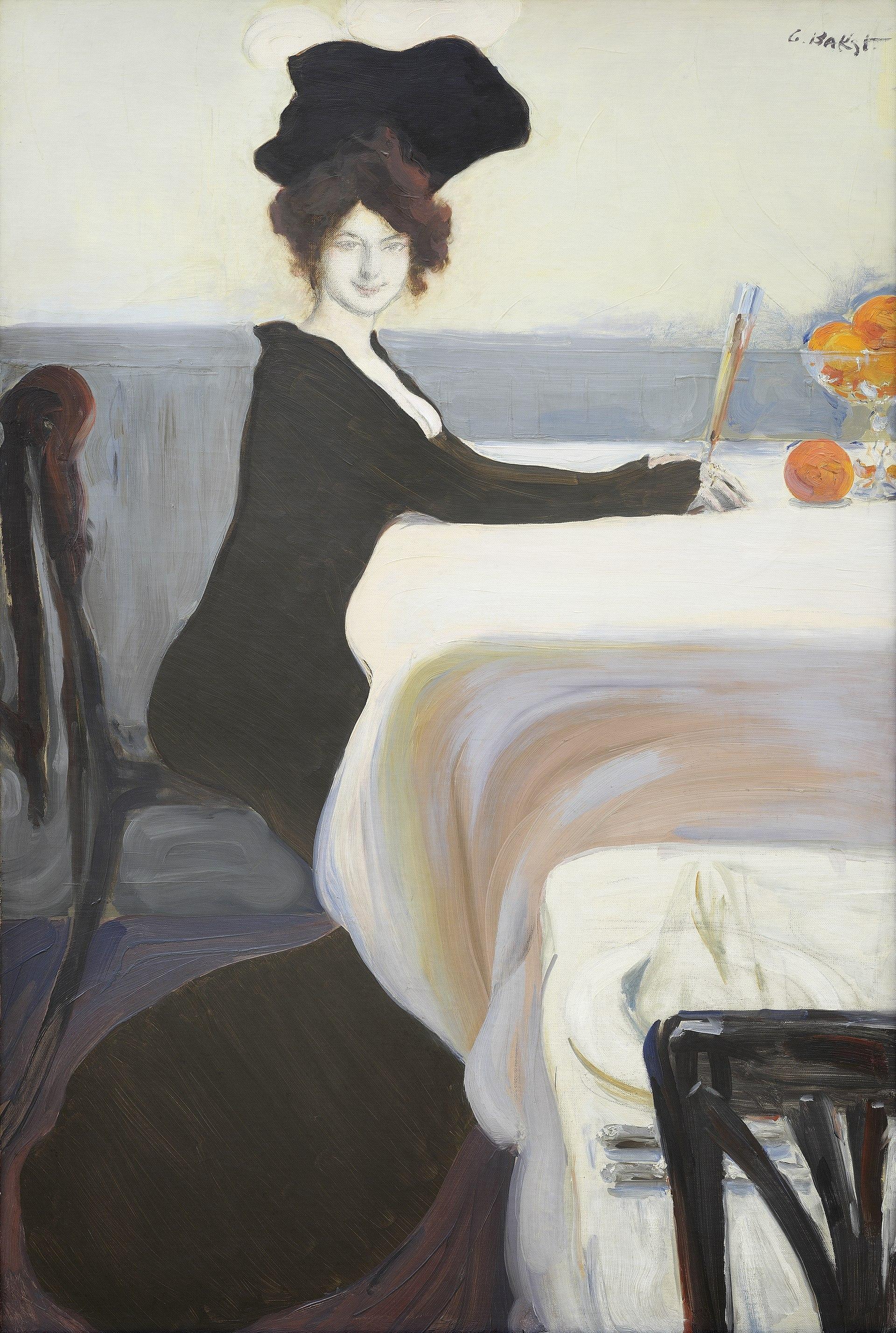 An elegant woman in black with an extravagant hat sits playfully tipping a flute of champagne towards a bowl of oranges. The tabes are covered with white, flowing tablecloths and the chairs are of black cane. She is alone